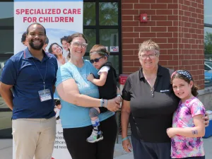 A DSCC Care Coordinator smiles next to a DSCC participant family of four. They’re all standing in front of DSCC’s Mokena office. The four-year-old DSCC participant sits in his mom’s arms. He is next to his seven-year-old sister and grandma.