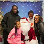 A DSCC participant family of four poses with Santa Claus in front of a festive holiday photo backdrop in DSCC's Mokena Regional Office