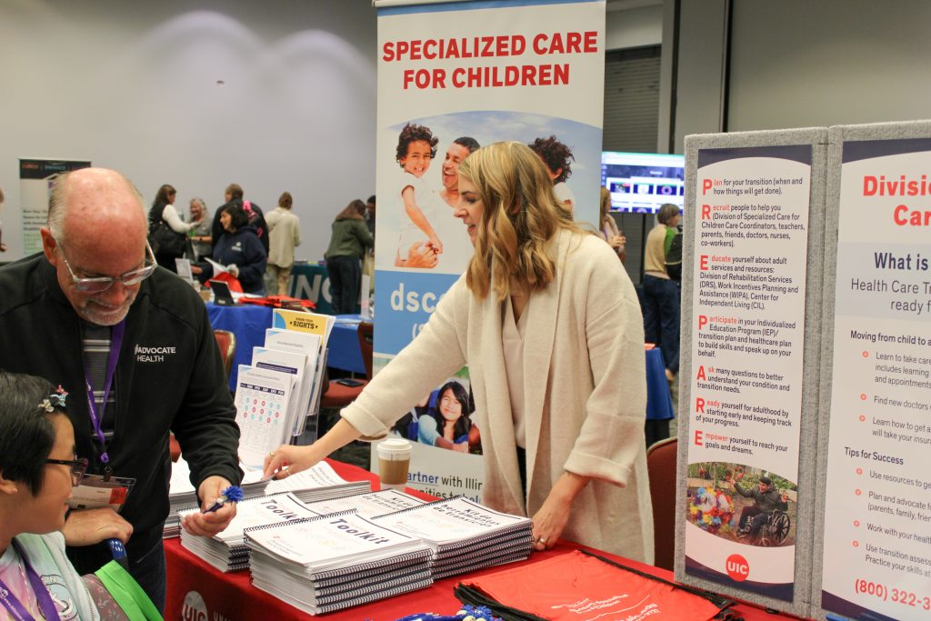 A DSCC team member hands out information at DSCC's booth at the Transition Conference Vendor Fair. A teenage DSCC participant and her father review the hand outs and take a promotional pen.