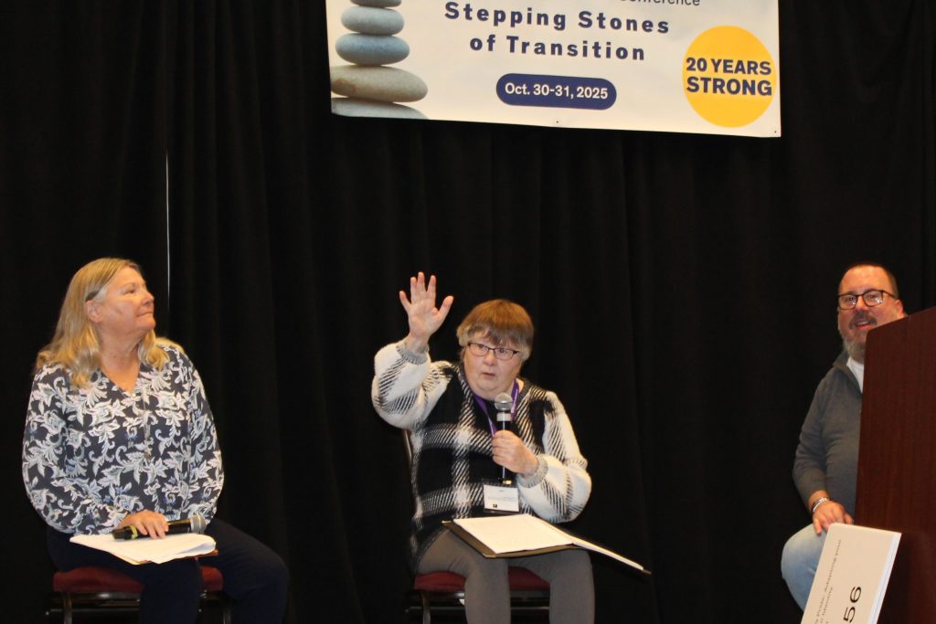 Keynote speaker Diana Braun speaks into a microphone on the conference stage. Two individuals sit next to her on stage. Diana raises her hand in the air to emphasize the point she is making.