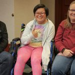 A family of three smiles together in the middle of a meeting room. The parents are on either side of their teenage daughter. She has a tie-dye shirt, colorful leggings, short hair, glasses and uses a wheelchair. She has a big smile and holds a thumbs-up.
