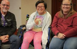 A family of three smiles together in the middle of a meeting room. The parents are on either side of their teenage daughter. She has a tie-dye shirt, colorful leggings, short hair, glasses and uses a wheelchair. She has a big smile and holds a thumbs-up.