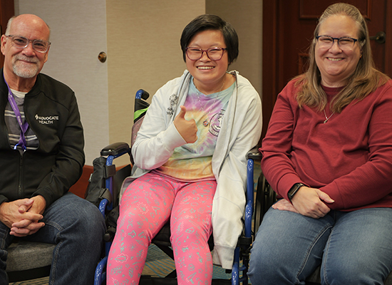 A family of three smiles together in the middle of a meeting room. The parents are on either side of their teenage daughter. She has a tie-dye shirt, colorful leggings, short hair, glasses and uses a wheelchair. She has a big smile and holds a thumbs-up.