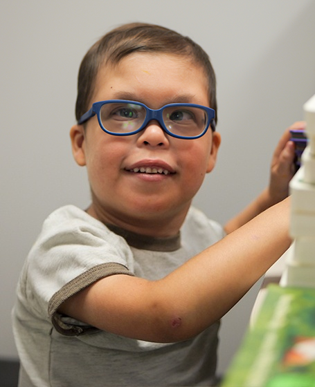 A young Division of Specialized Care for Children participant wearing glasses looks up and smiles at the camera as he plays