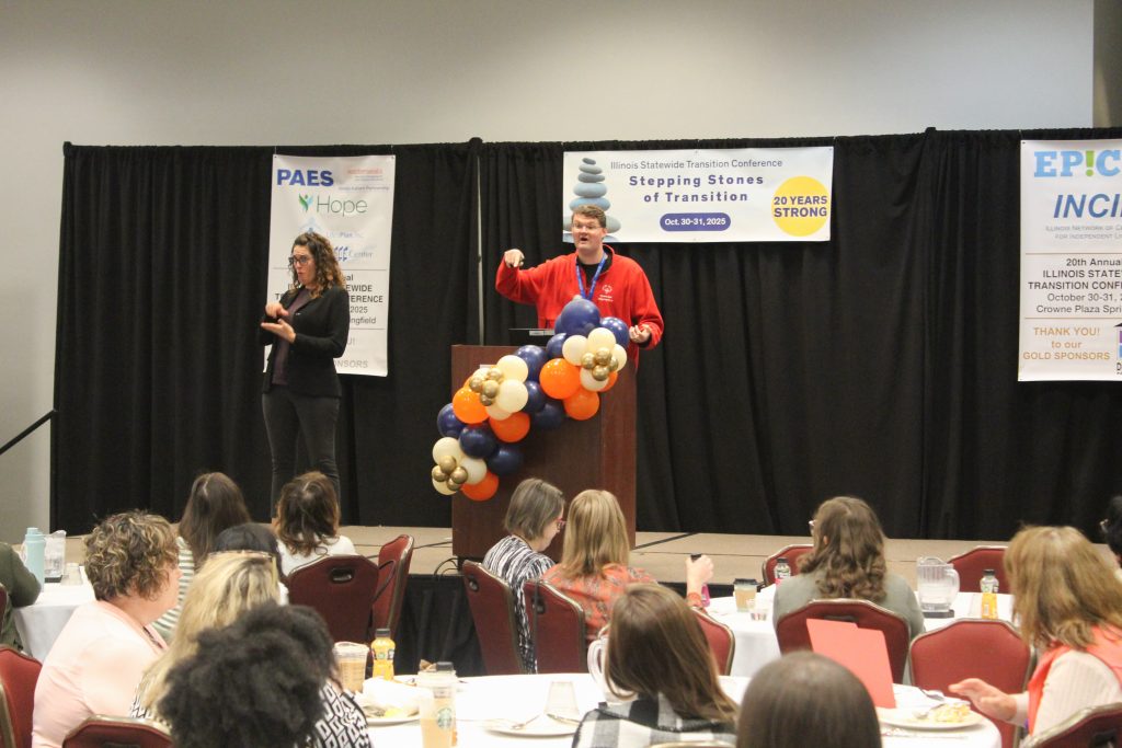 Keynote presenter Taylor Sweeting speaks into a microphone enthusiastically on the conference stage. An American Sign Language (ASL) interpreter signs next to him. The conference room is full with individuals sitting at dining tables.