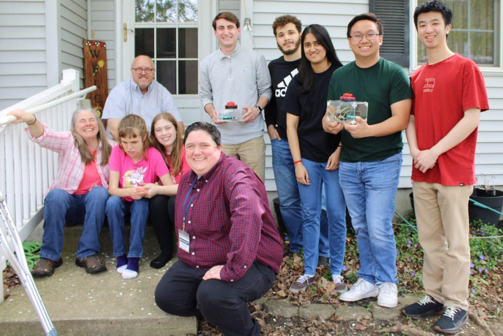 The MedLaunch team hold the SeizureSense Safety Alert System next to Makaelyn, a young adult, her parents, caregiver and care coordinator. They smile outside the family home.