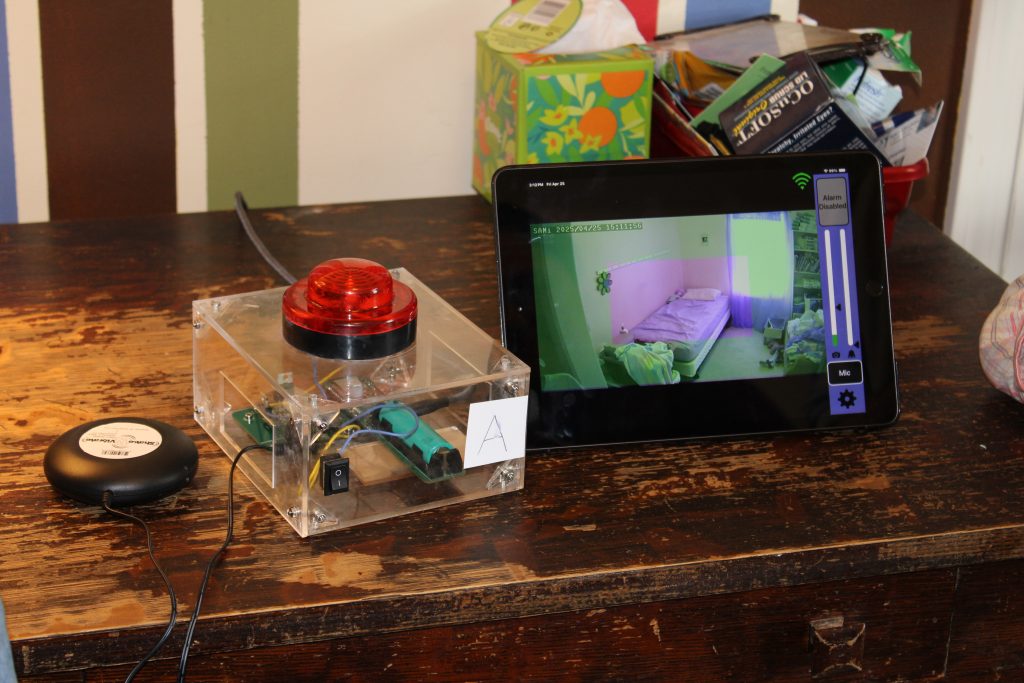 Close up of the SeizureSense Safety Alert System on a bedside table. The alert system has a large red button on top of a clear box with visible wires. There is a tablet next to the alert system which shows the monitored bedroom.