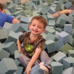 A young Division of Specialized Care for Children participant with a limb difference smiles as he plays in the foam block pit of the Sensory Station at the Illinois State Fair