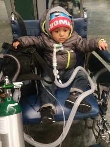 DSCC participant Dominic Garvin as a toddler, seated in a wheelchair, wearing a winter coat and hat, with a tracheostomy connected to breathing equipment and an oxygen tank mounted beside the wheelchair.