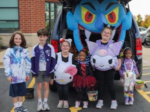 A van trunk decorated with a Halloween evil genie blow up. A group of six youth stand in front of the trunk in their costumes smiling for the camera.