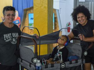 DSCC participant Dominic Garvin sits inside his stroller wagon while his mom, Aurea, and his grandmother stand beside him and smile